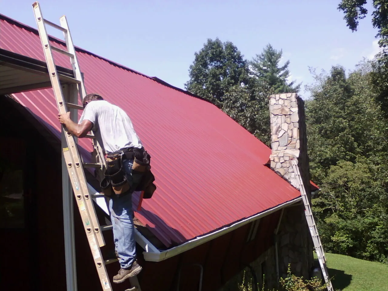 Roofer on a ladder beside a red metal roof and stone chimney — Soffit & Fascia Repair by Roofing Brothers in Kesley